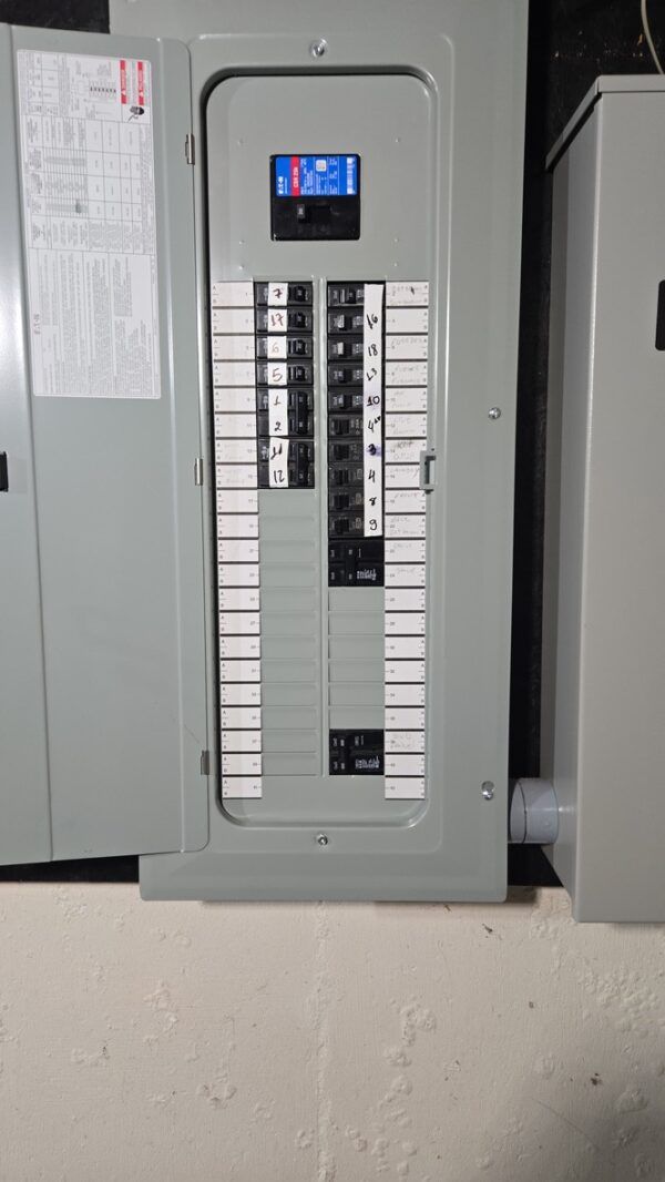 Electrical panel with multiple circuit breakers, mounted on a wall. Gray box, partially open, with labels and switches.