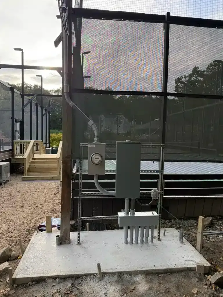 Electrical box and meter mounted on concrete pad near a sports court, with wire conduits.