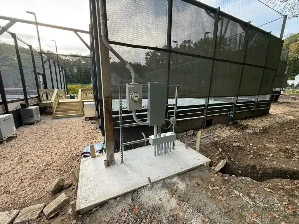 Electrical box and conduit installed on a concrete pad near a shaded enclosure with gravel ground.