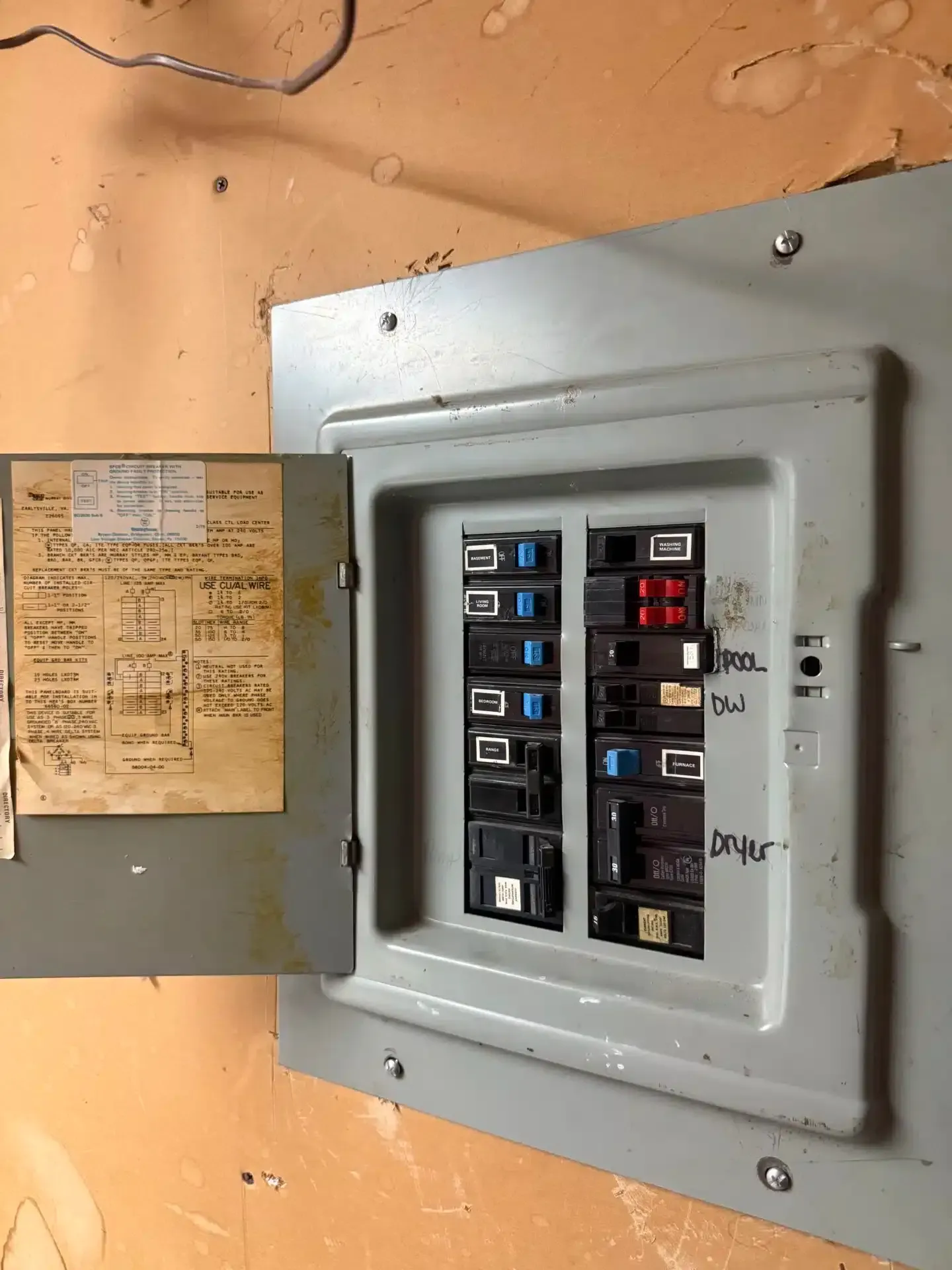 Electrical circuit breaker panel with open door, showing labeled switches against an orange wall.