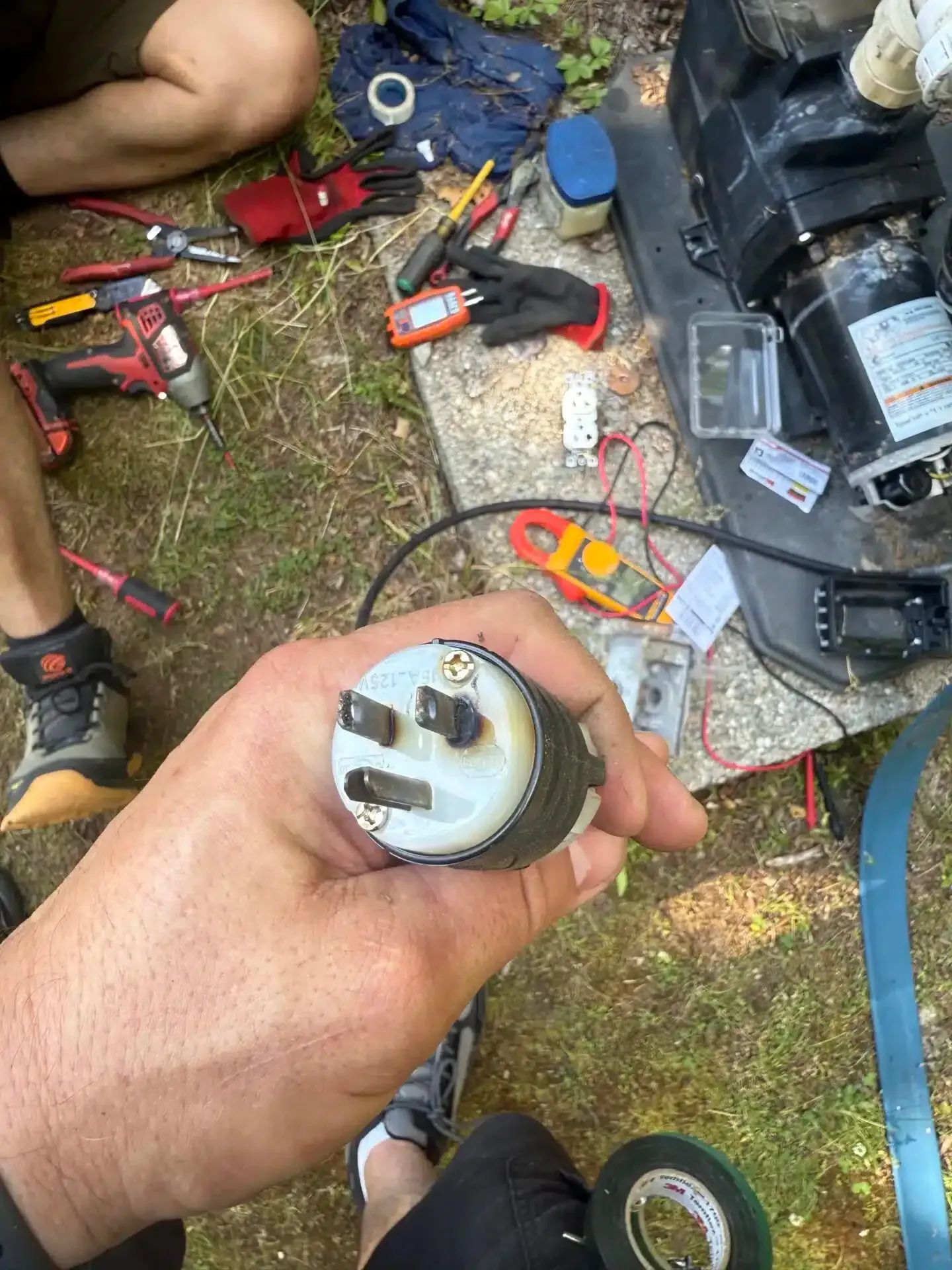 Hand holding a three-prong electrical plug. Tools and equipment are scattered on the ground around it.