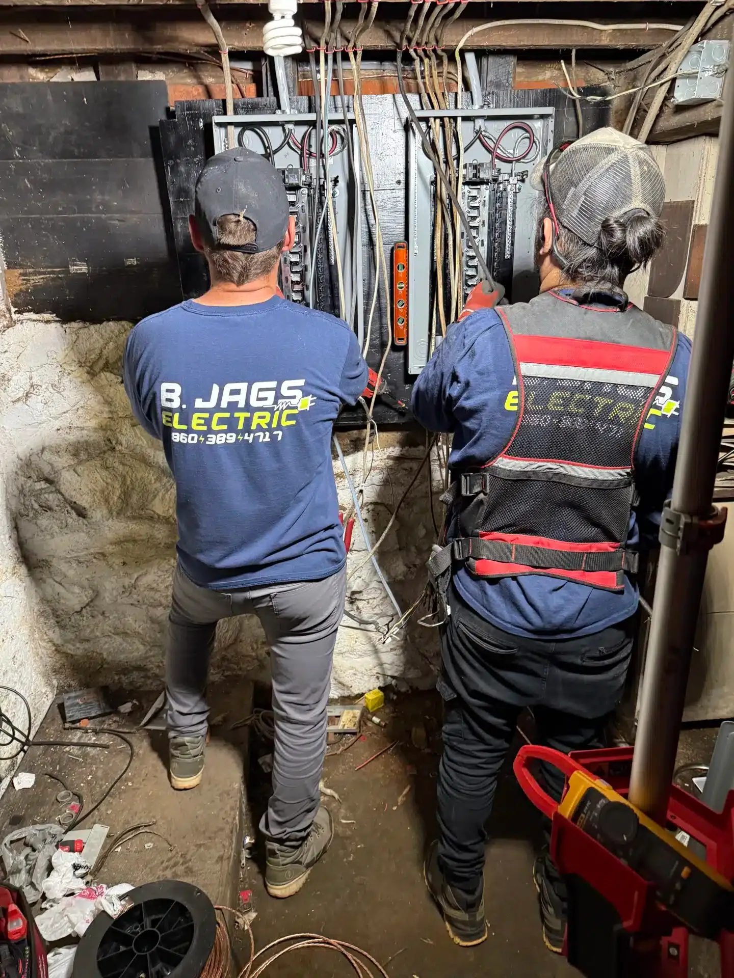 Two electricians working on electrical panels in a basement. One is wearing a blue shirt, the other a safety vest.