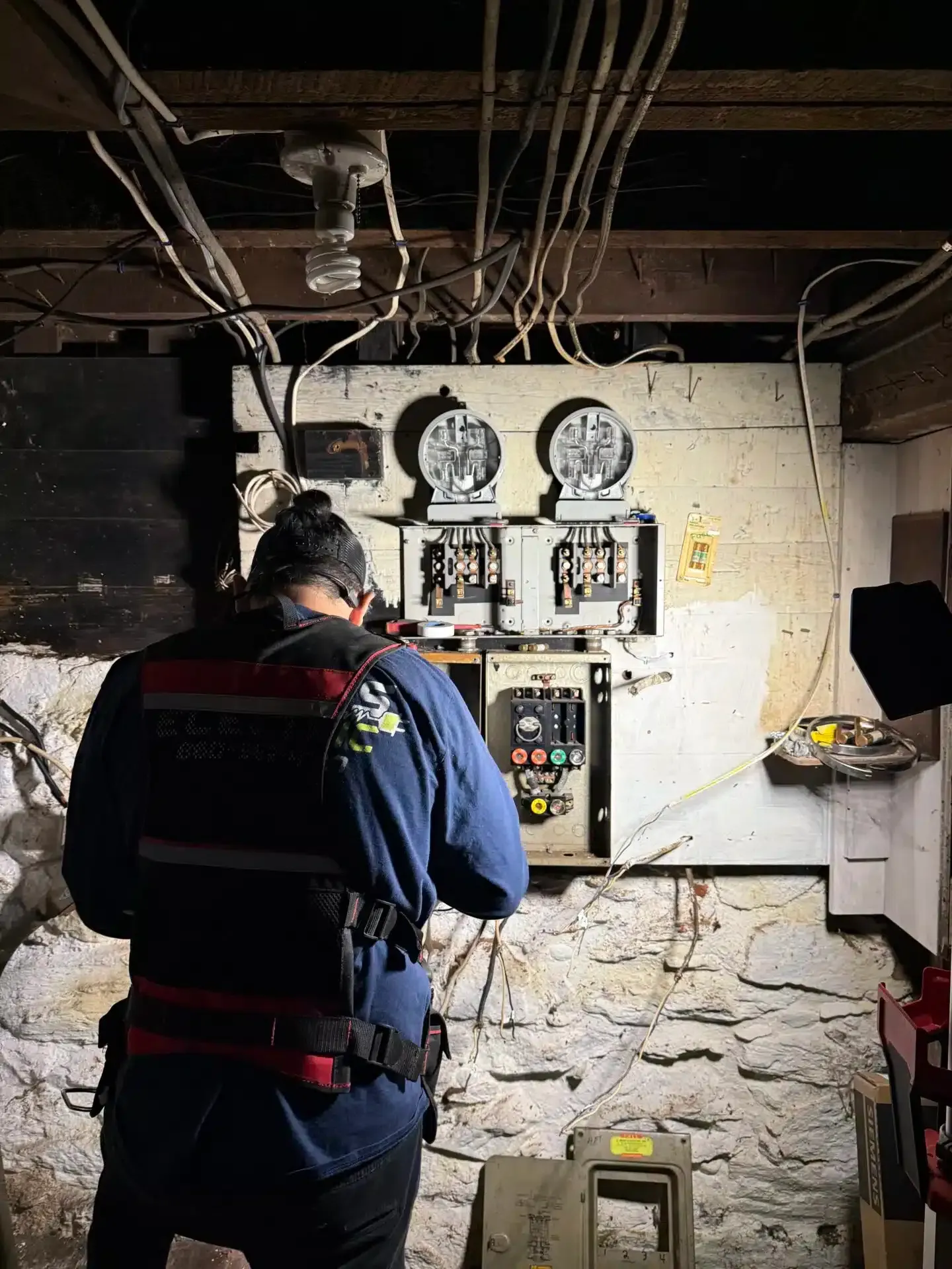 Electrician working on electrical panel in a basement. Wires, meters, and tools visible.