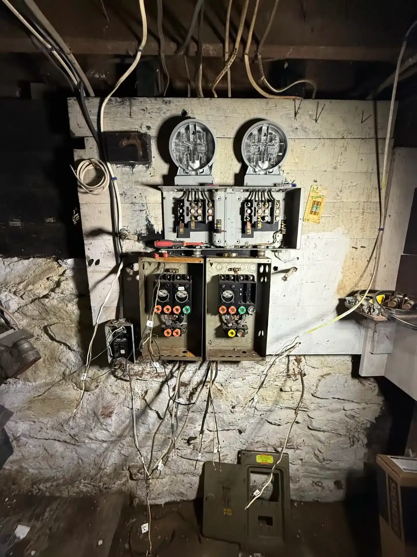 Old electrical panel with meters and circuit breakers on a rough, white brick wall in a basement.