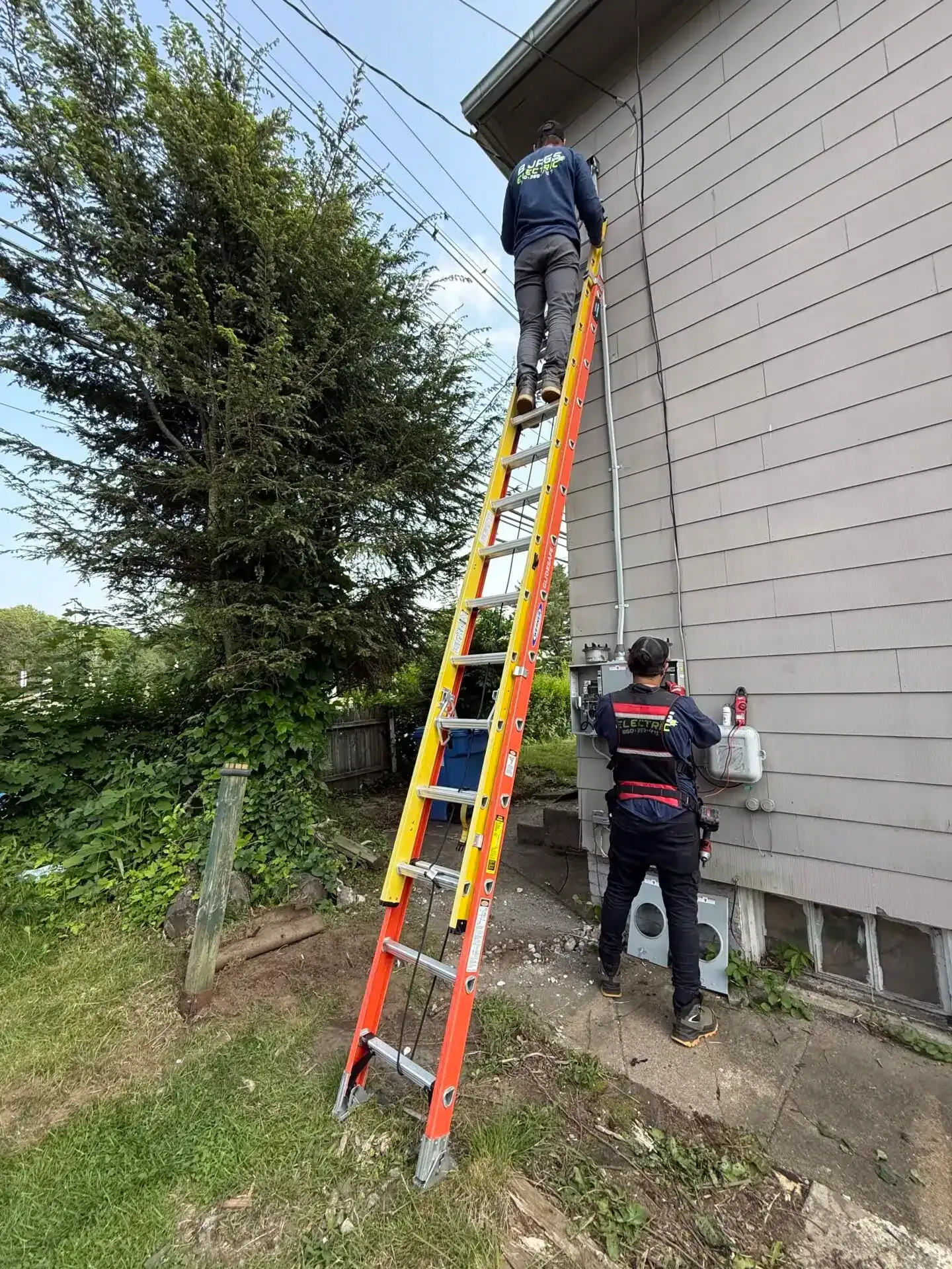 Two workers on a tall ladder, one at the roofline, the other near electrical work on the house exterior.