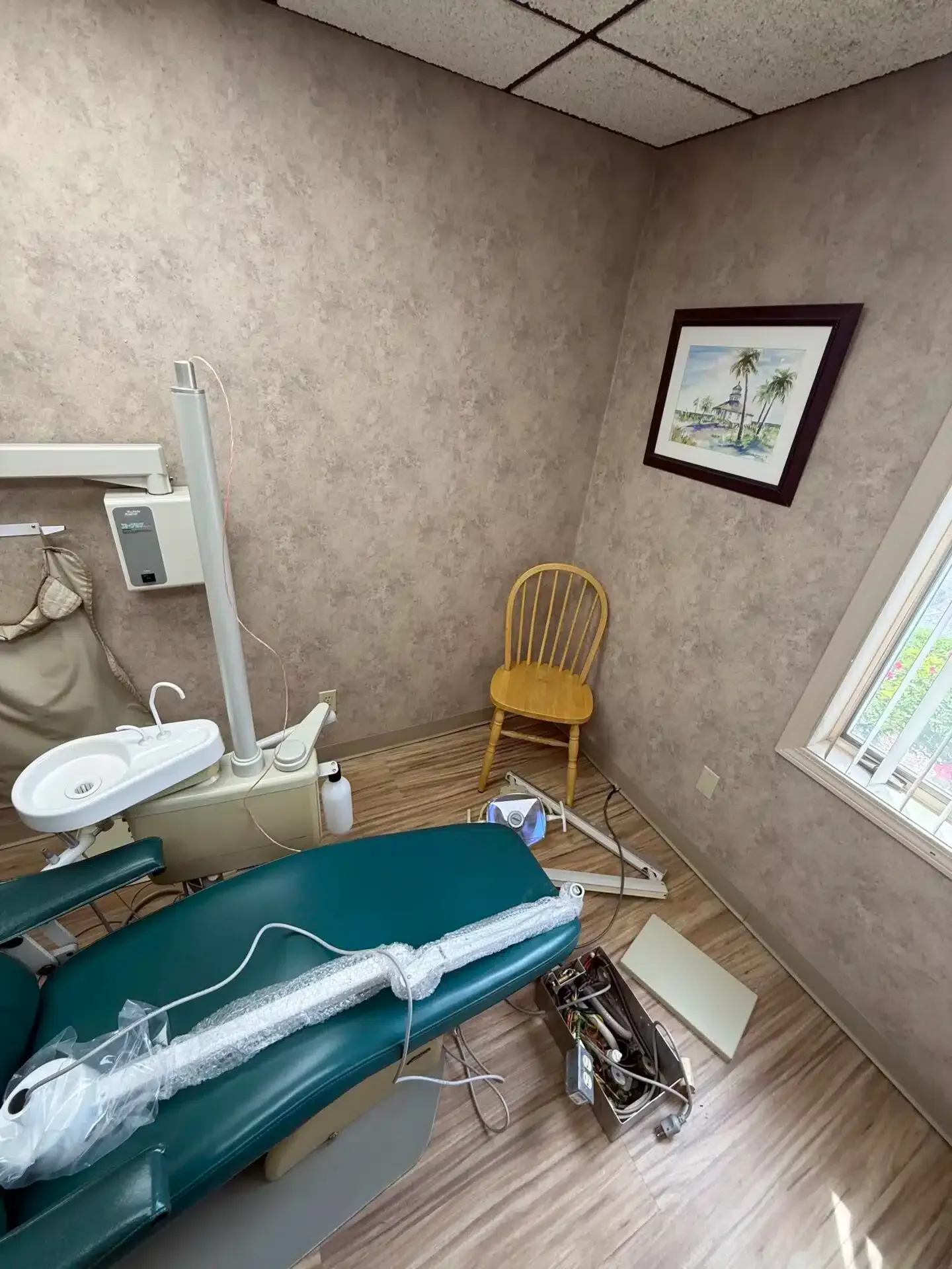 Dental office interior; dental chair, wash basin, chair, artwork on wall, window, brown/beige walls.