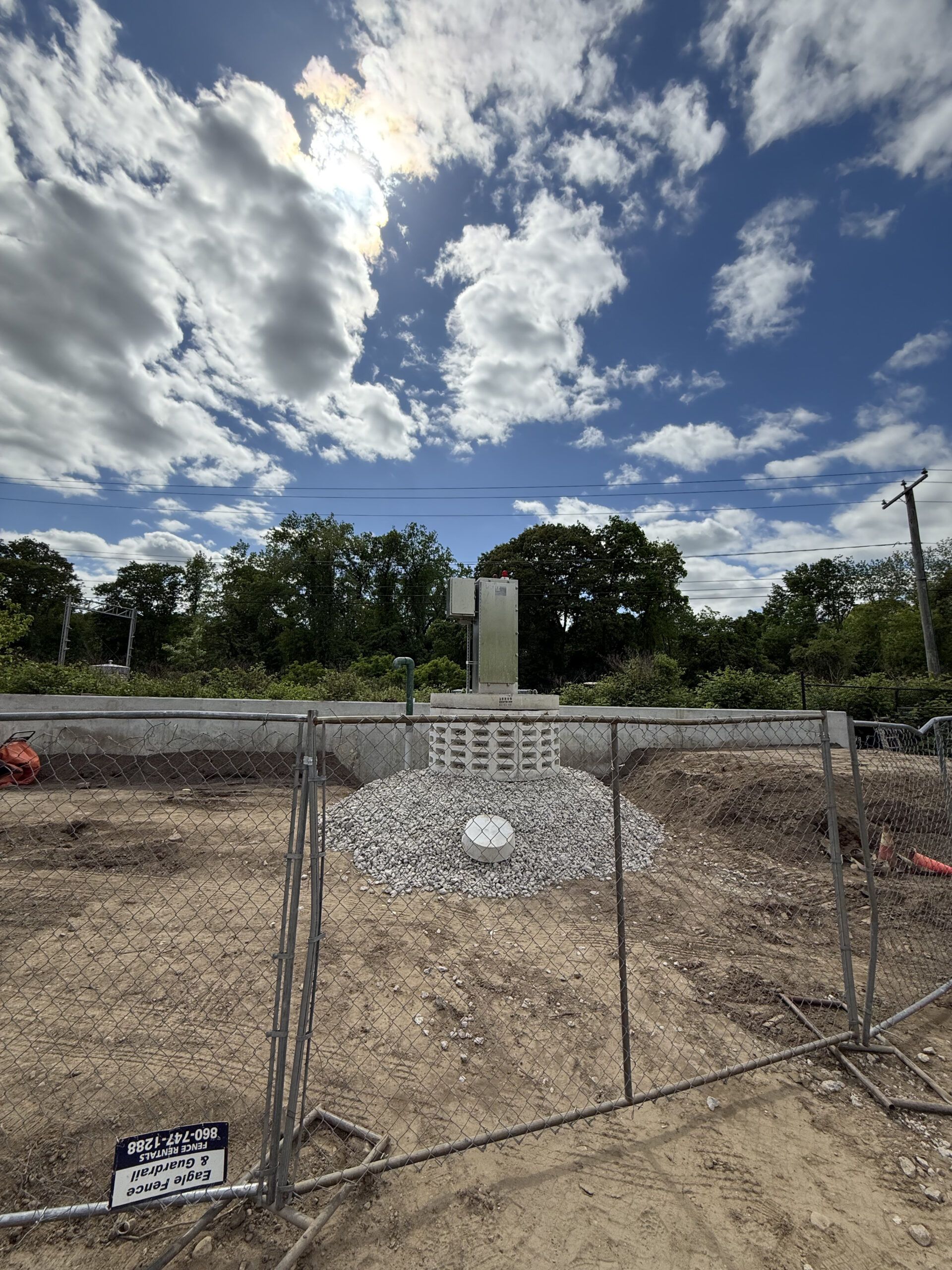 Construction site with a metal structure against a cloudy blue sky. A chain link fence is in front.