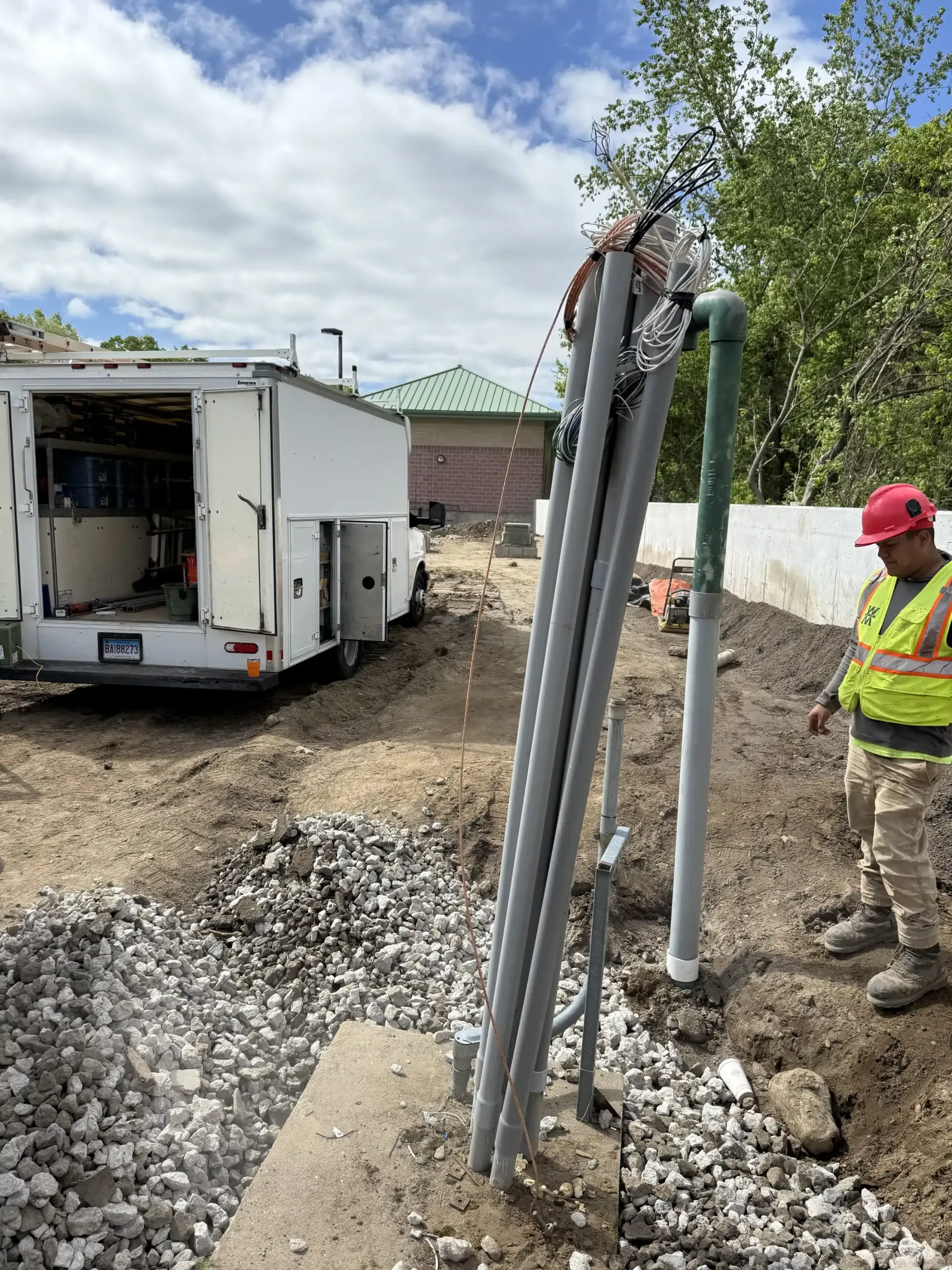 Construction worker near pipes, trailer, and ground excavation. Green and gray pipes; gravel. Sunny, outdoor setting.