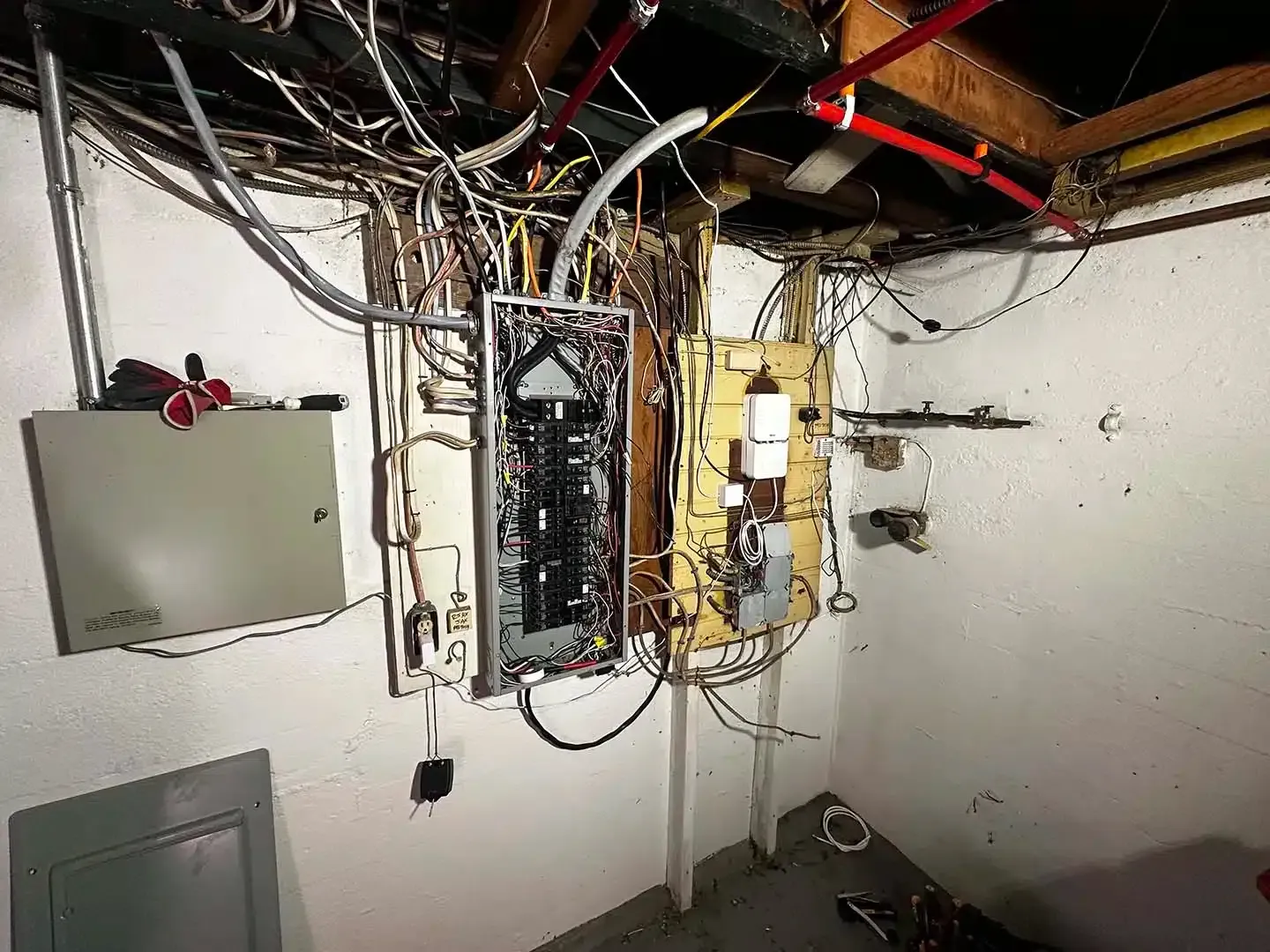 Electrical panels and wiring in a basement. Wires are tangled and messy; red water pipes overhead.