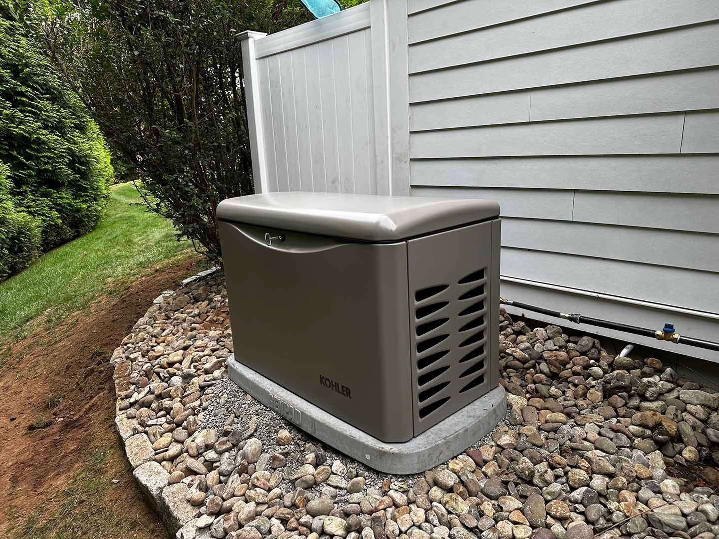 Tan standby generator on a gravel base beside a white fence and house with gray siding.