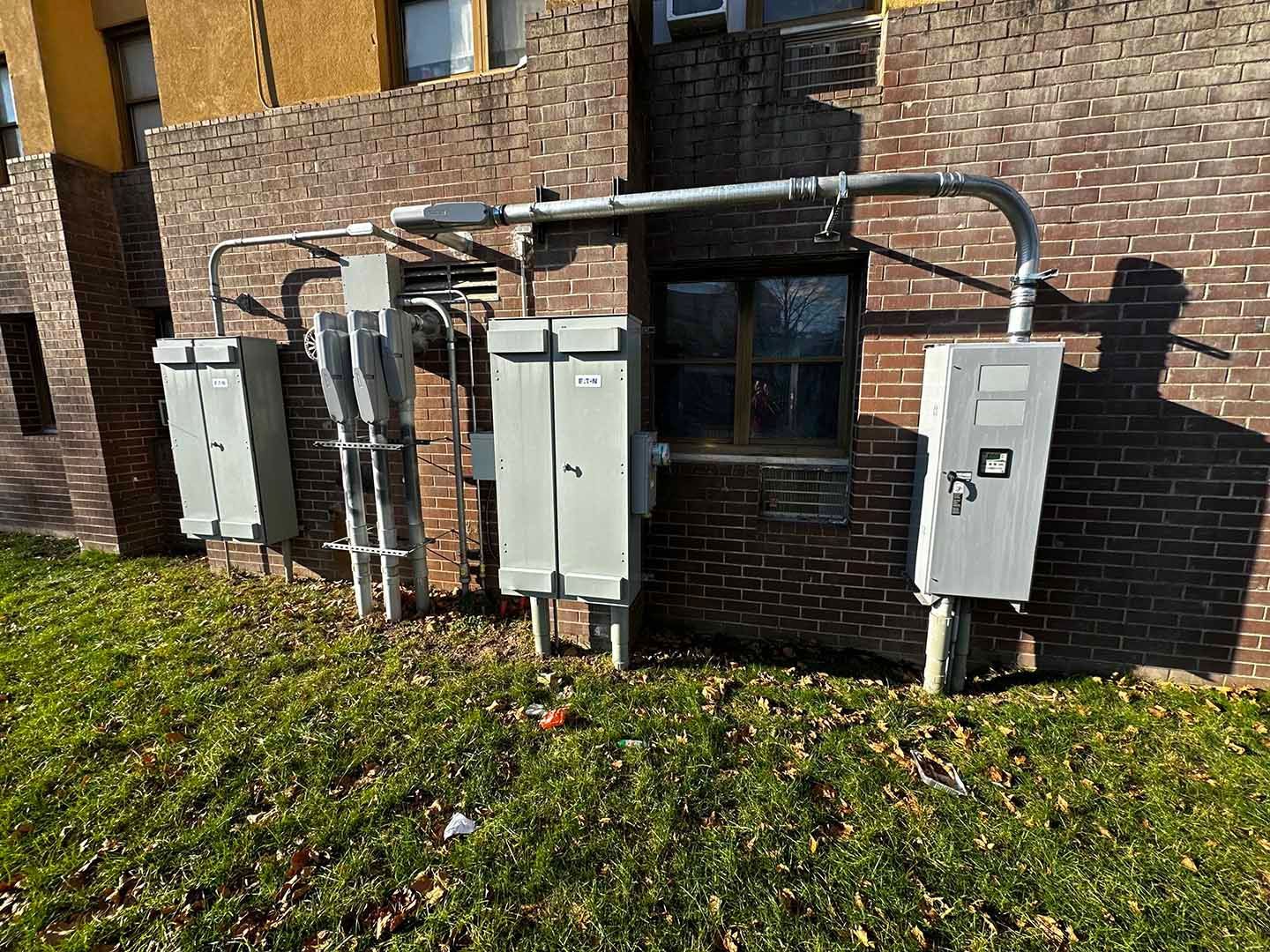 Electrical equipment on brick building exterior; grey metal boxes and conduits on grassy ground.