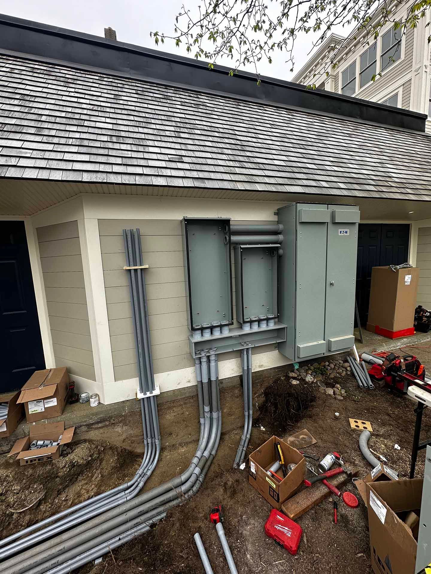 Exterior electrical work on a building: conduits, panels, and a large gray box on a tan wall.