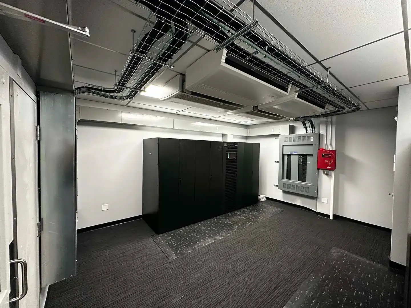 Server room with black server racks, electrical panel, and cables on the ceiling.