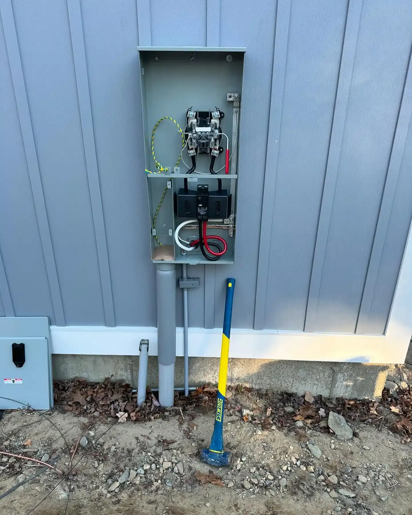 Electrical panel mounted on a gray building exterior, with conduits and a hammer nearby.