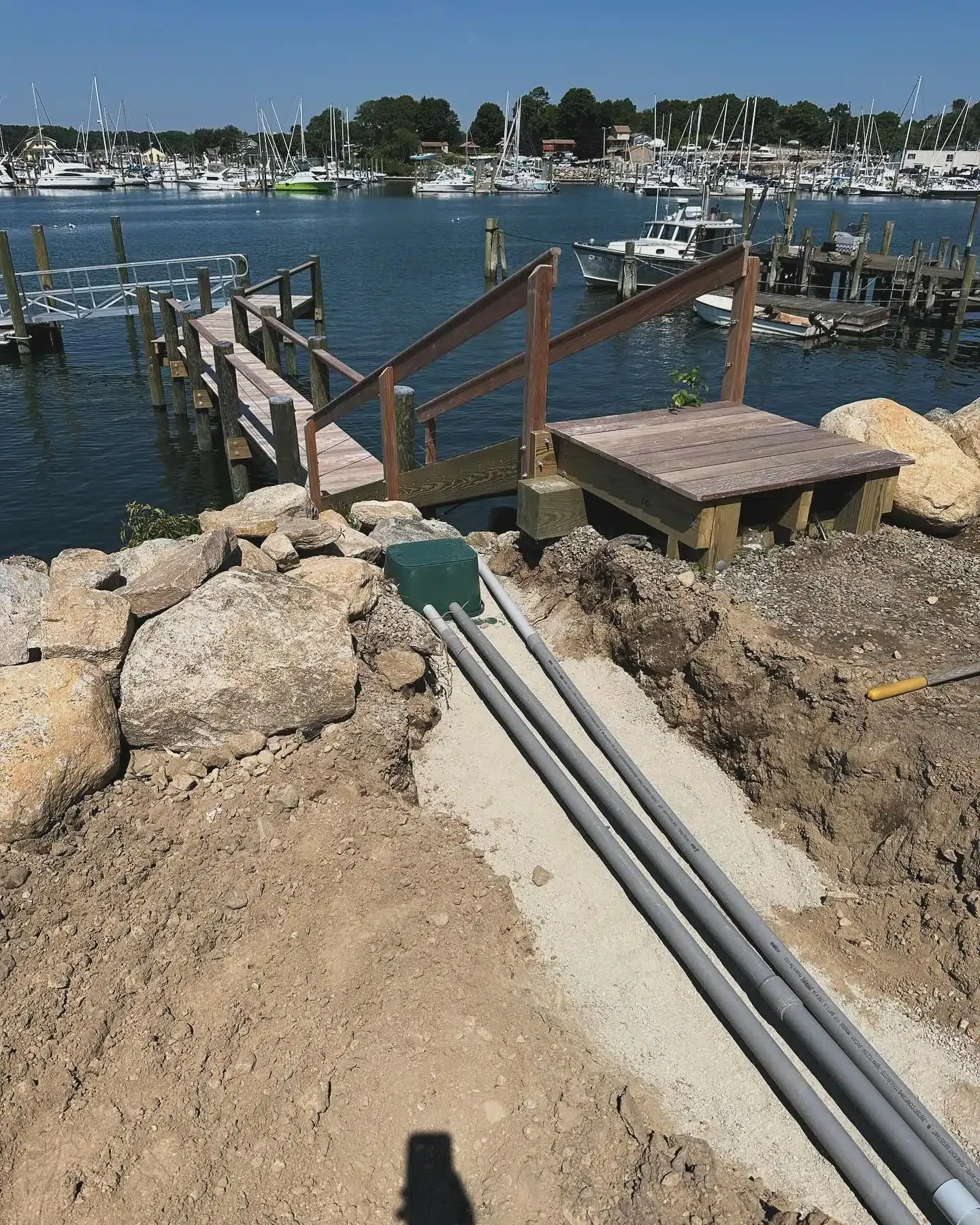Construction site near a marina. Conduit pipes run from a green box towards a wooden pier, next to dirt and rocks.