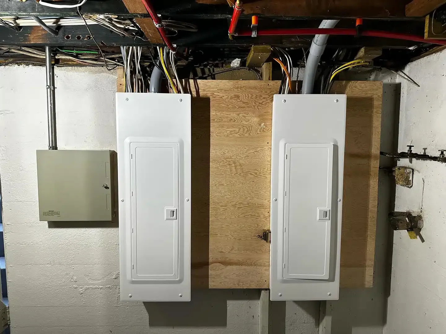 Two white electrical panels mounted on plywood in a basement. Wires and conduit are visible.
