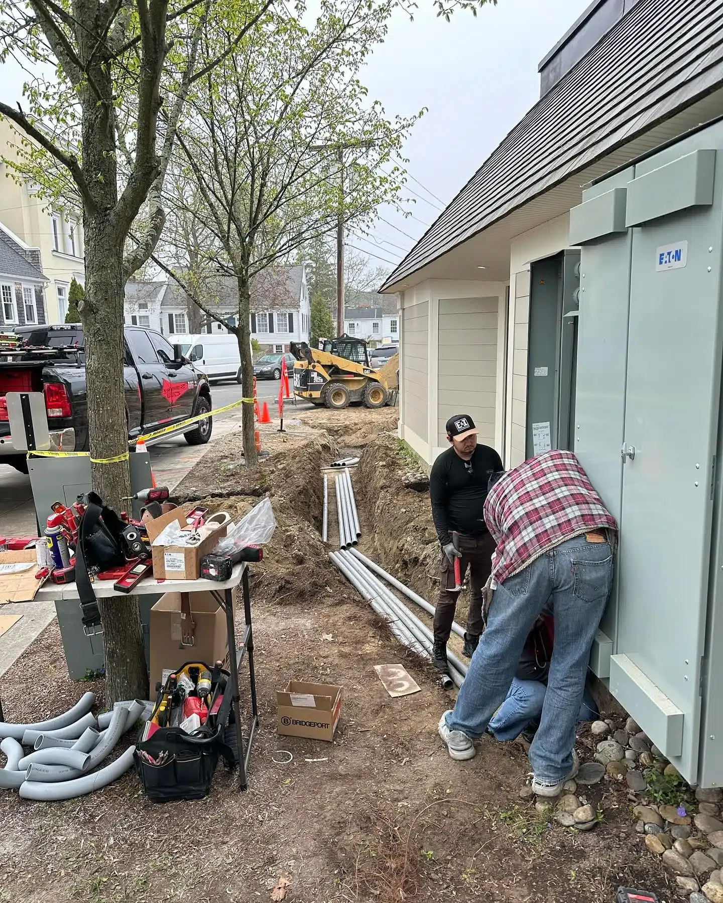 Construction workers installing electrical equipment next to a building. Tools and supplies are visible on the ground.