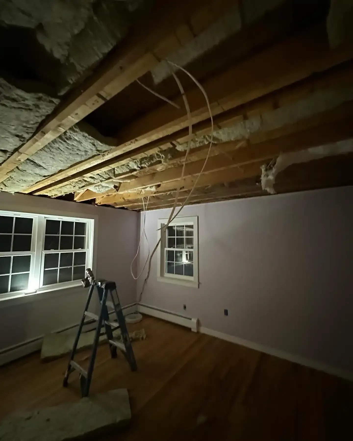 Room under construction with exposed ceiling beams and electrical wiring. A ladder stands by a window.