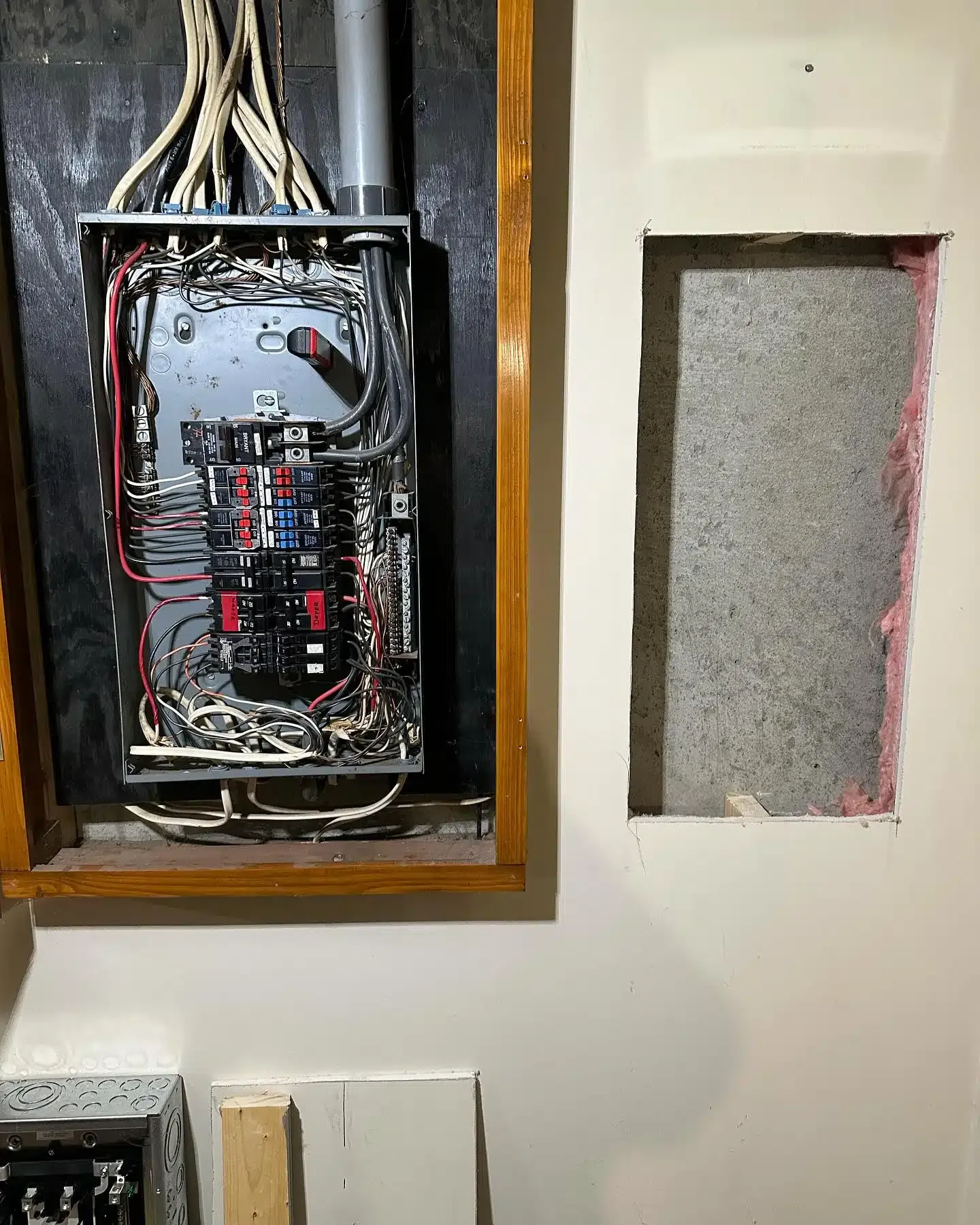 Electrical panel open, wires visible, next to a wall with a rectangular hole.
