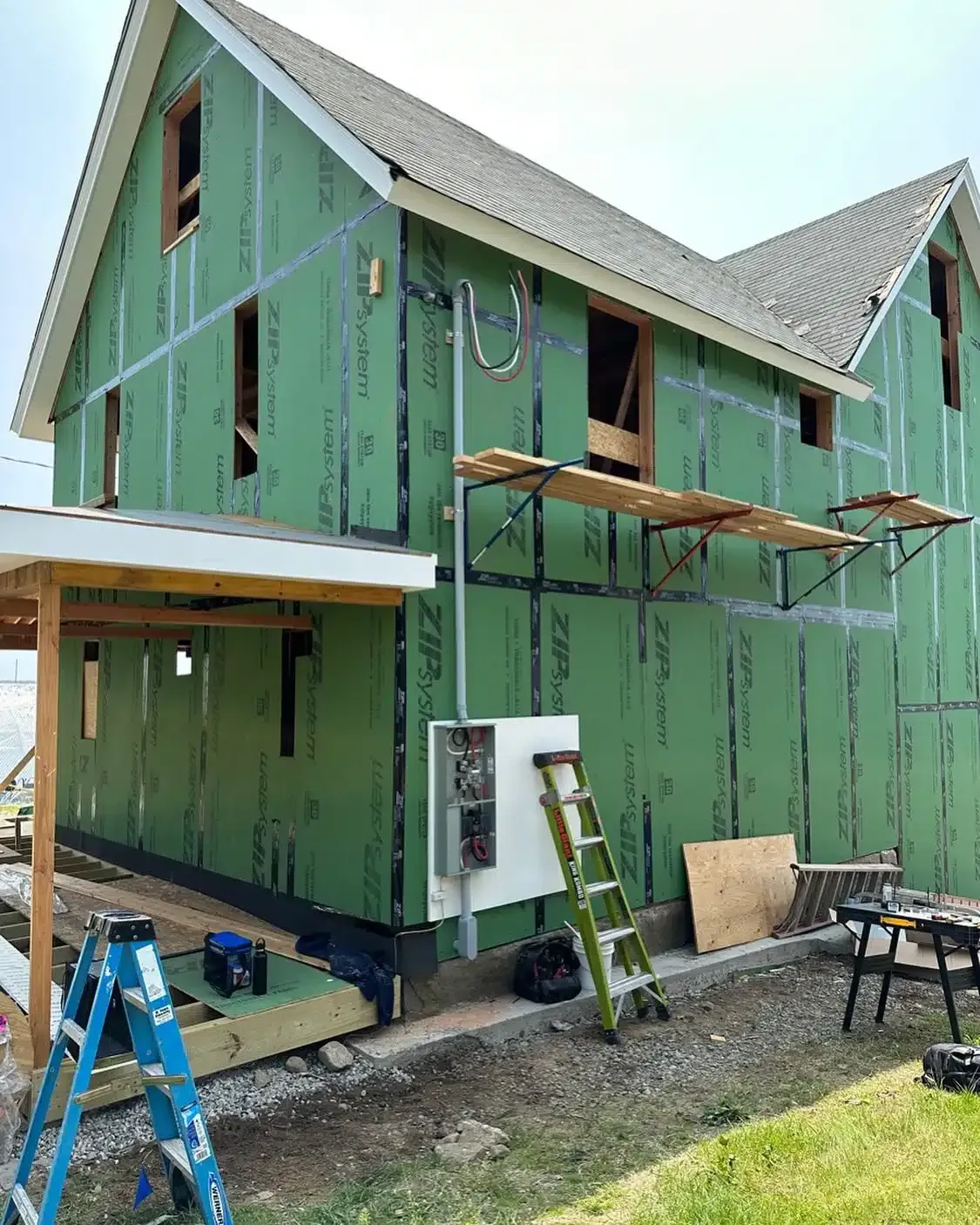 Exterior view of a house under construction; green sheathing, electrical panel, scaffolding, and ladder are visible.