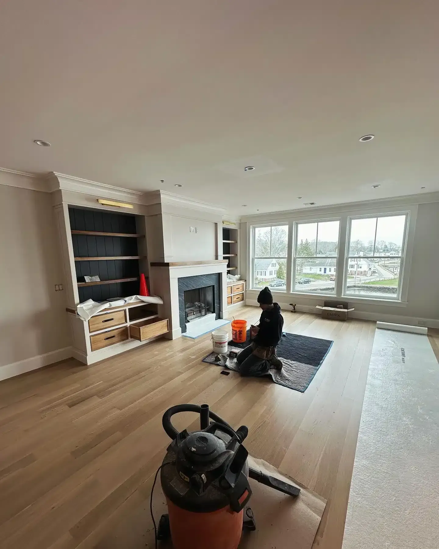 A person kneels on a rug in a room, working near a fireplace and bookshelves. An orange vacuum cleaner sits in the foreground.