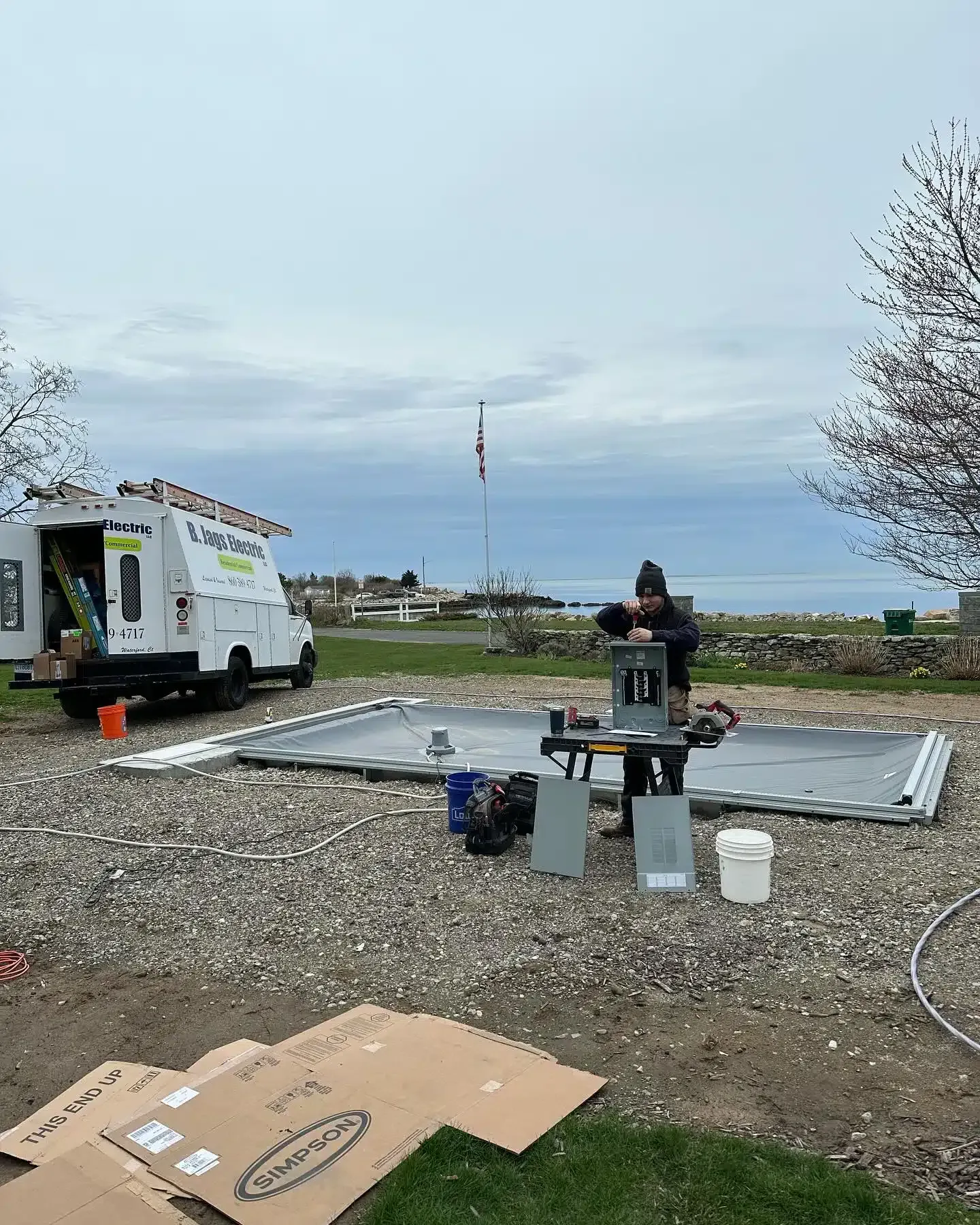 A person works on a metal structure outside, near a van, water, and cloudy sky.