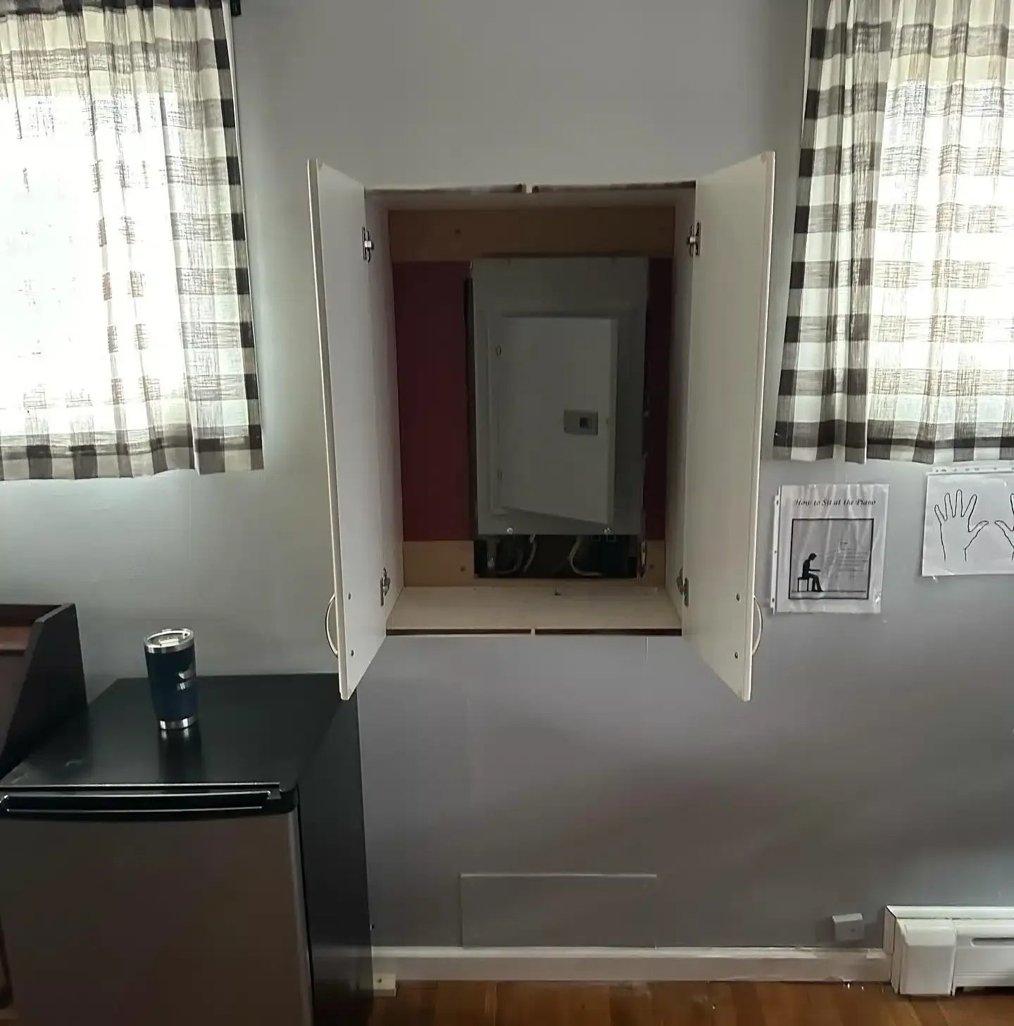 Cabinet doors open to reveal an electrical panel. Curtains and mini fridge are in the room.