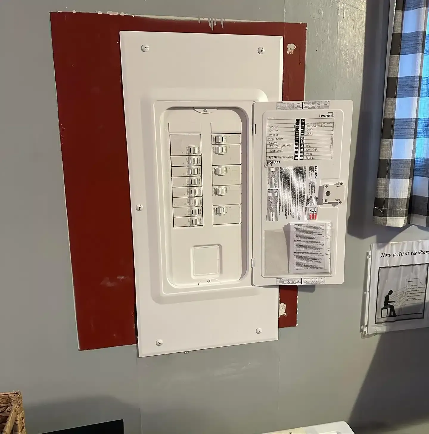 White electrical breaker box mounted on a red backing against a gray wall. Open panel door.