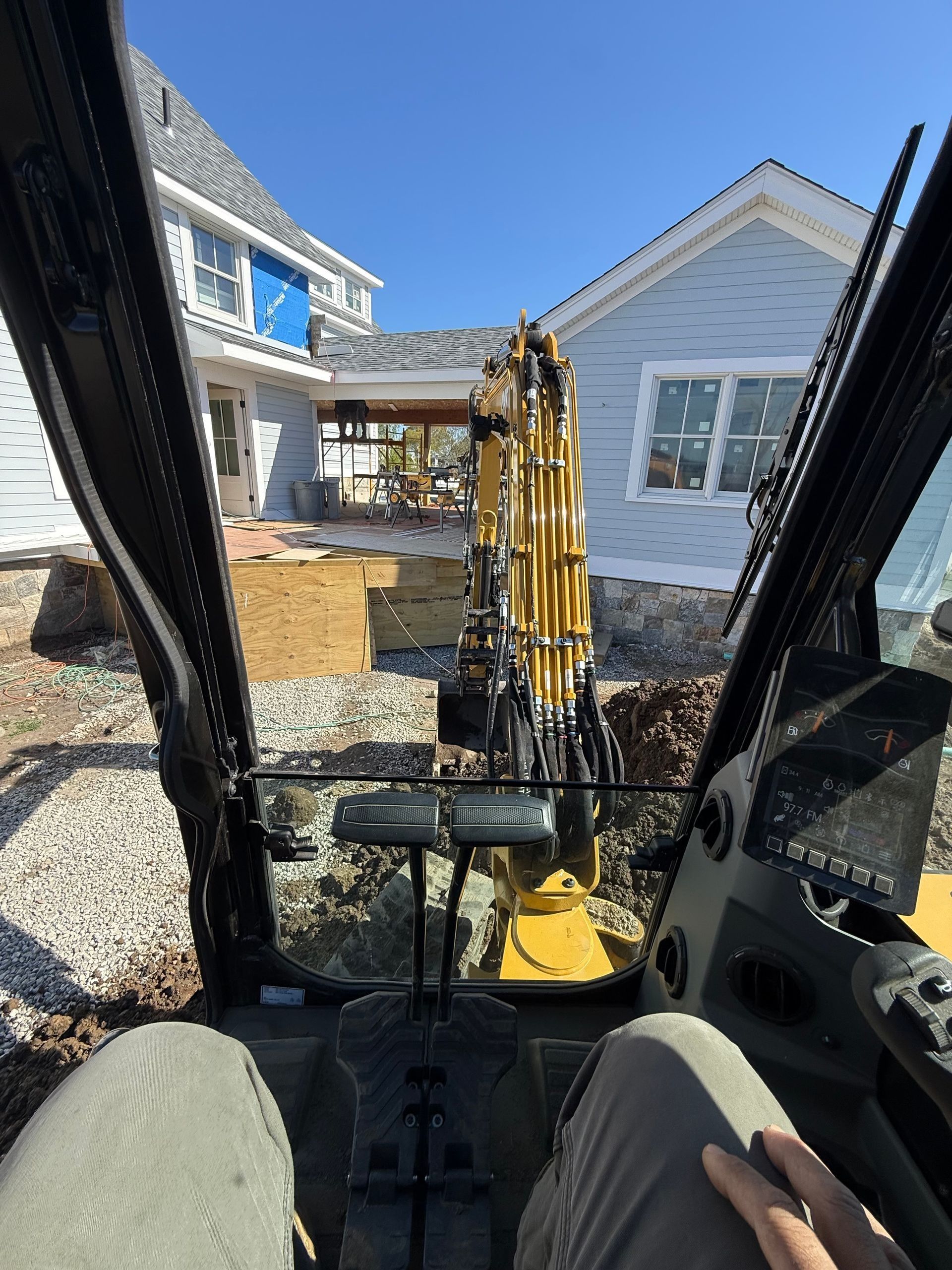 Excavator cab view of a yellow machine digging beside houses on a sunny day