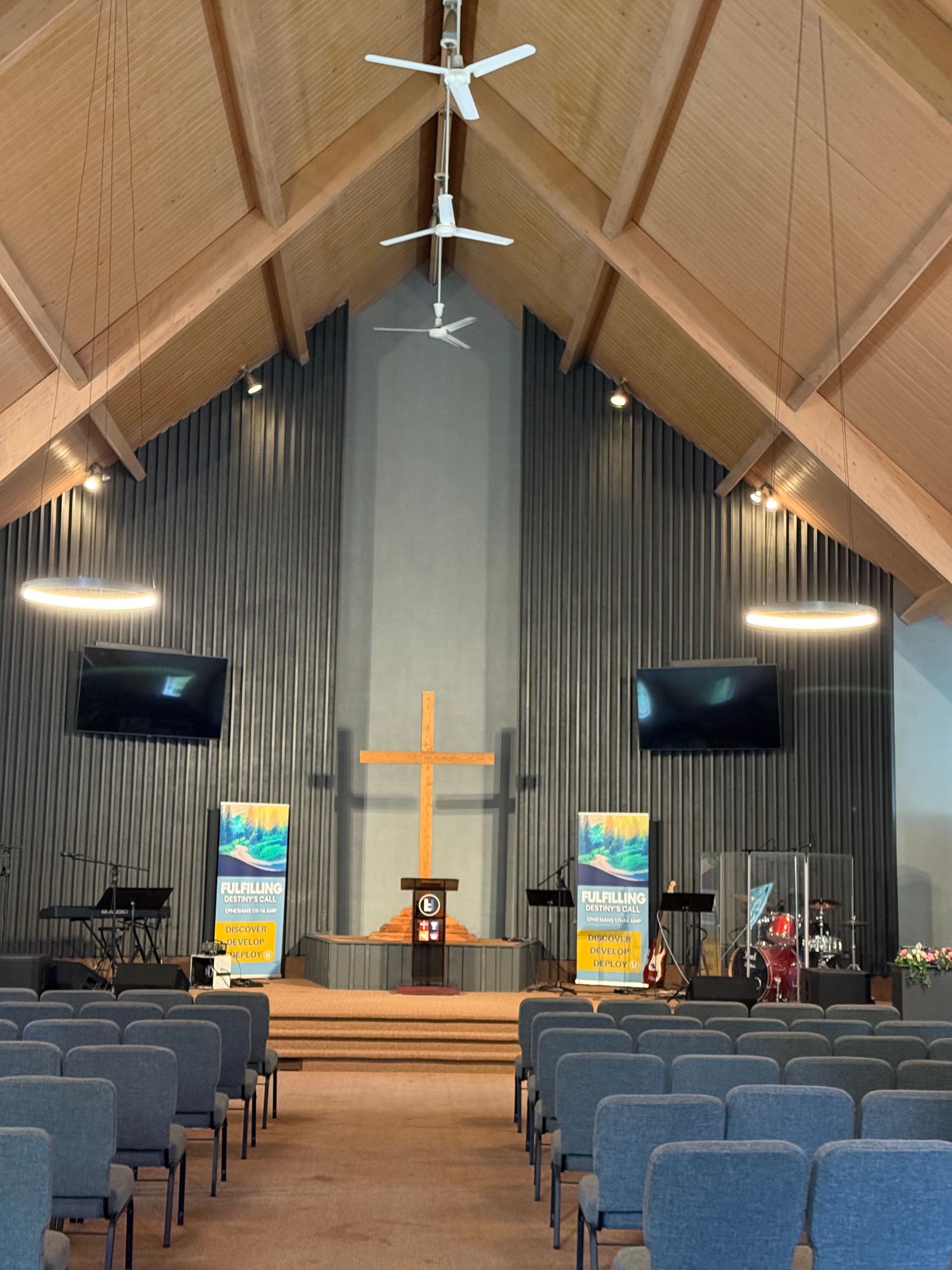 Church sanctuary with a wooden cross at the altar, blue chairs, and hanging monitors under a vaulted ceiling