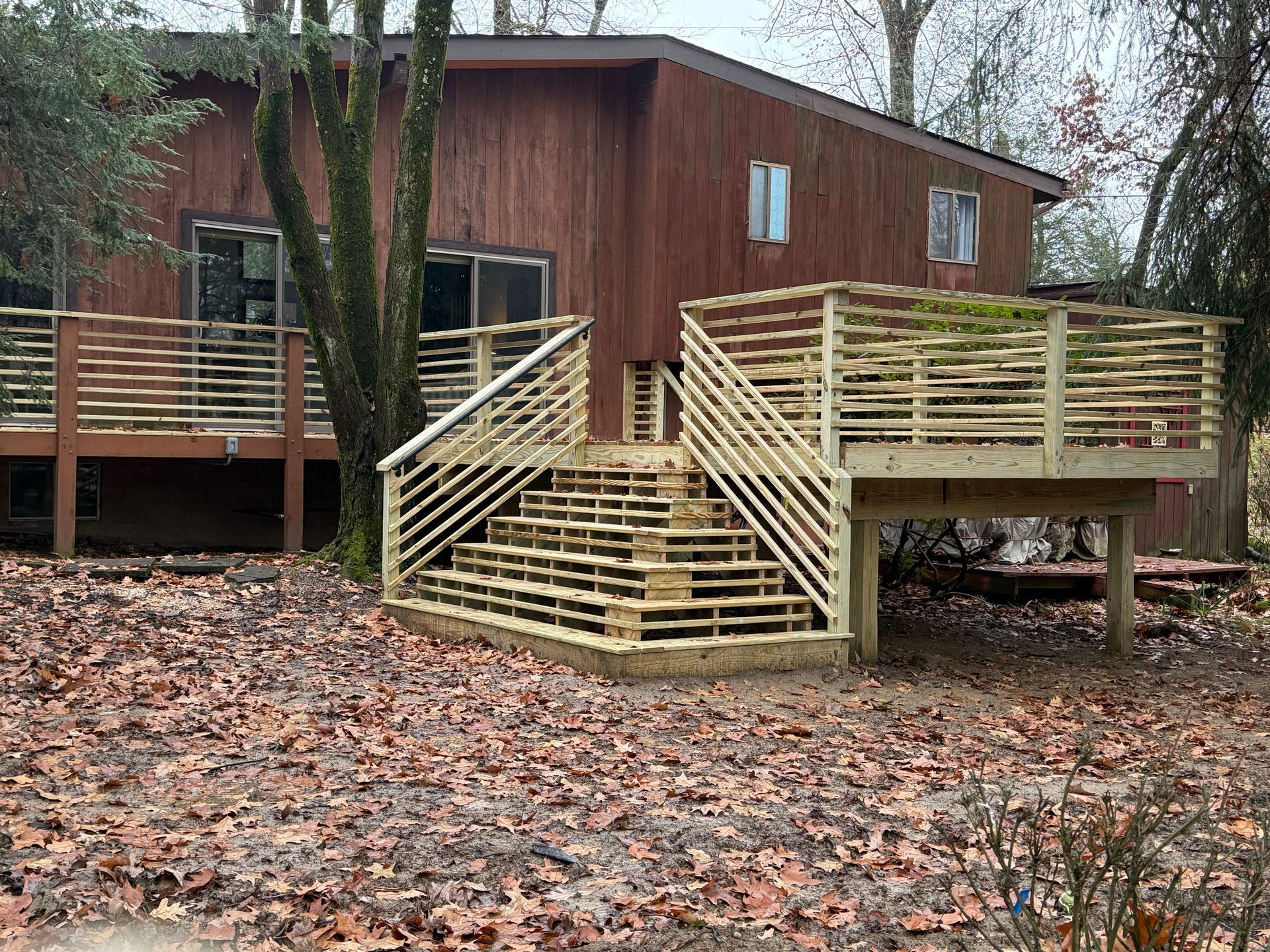 Wooden deck with stairs leading to a brown house; dry leaves on the ground.