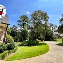 A stone house and circular concrete driveway fronted by a lush green lawn and mature trees under a clear blue sky.