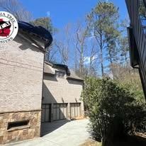 A beige brick house with a multi-car garage, concrete driveway, and tall trees under a bright blue sky.