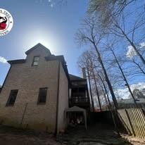 A tan brick house stands under a bright blue sky, next to tall, leafless trees and a wooden fence.