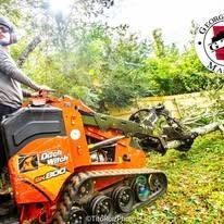 A person operating an orange Ditch Witch SK800 track loader to move a fallen tree in a wooded area.