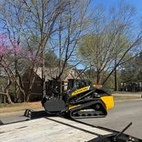 A yellow and black New Holland compact track loader sits on a flatbed trailer on a street with trees in the background.