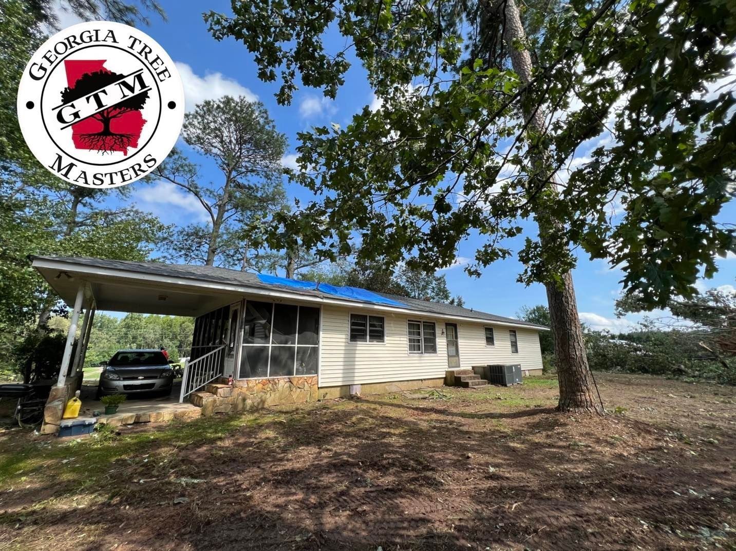 A single-story house with a blue tarp on its roof and a carport, surrounded by trees on a sunny day.