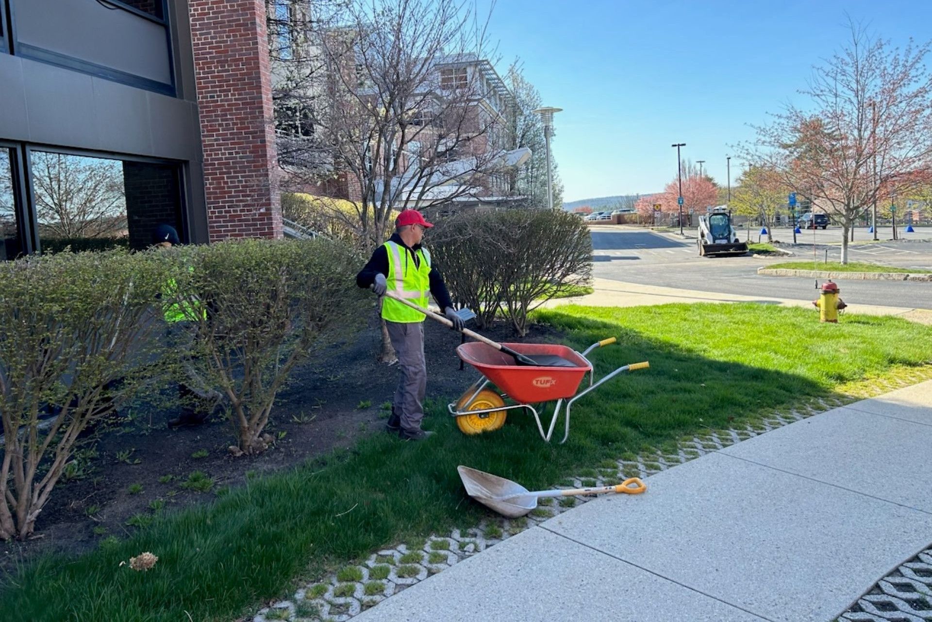 Person in safety vest and hat rakes near a wheelbarrow and shovel in a grassy area outside a building.