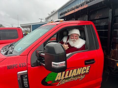 Santa giving a thumbs up from the cab of a red Alliance Landscaping truck.