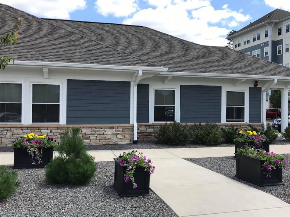 Building with blue siding, stone base, white trim, and a gray roof. Flower planters line the walkway.