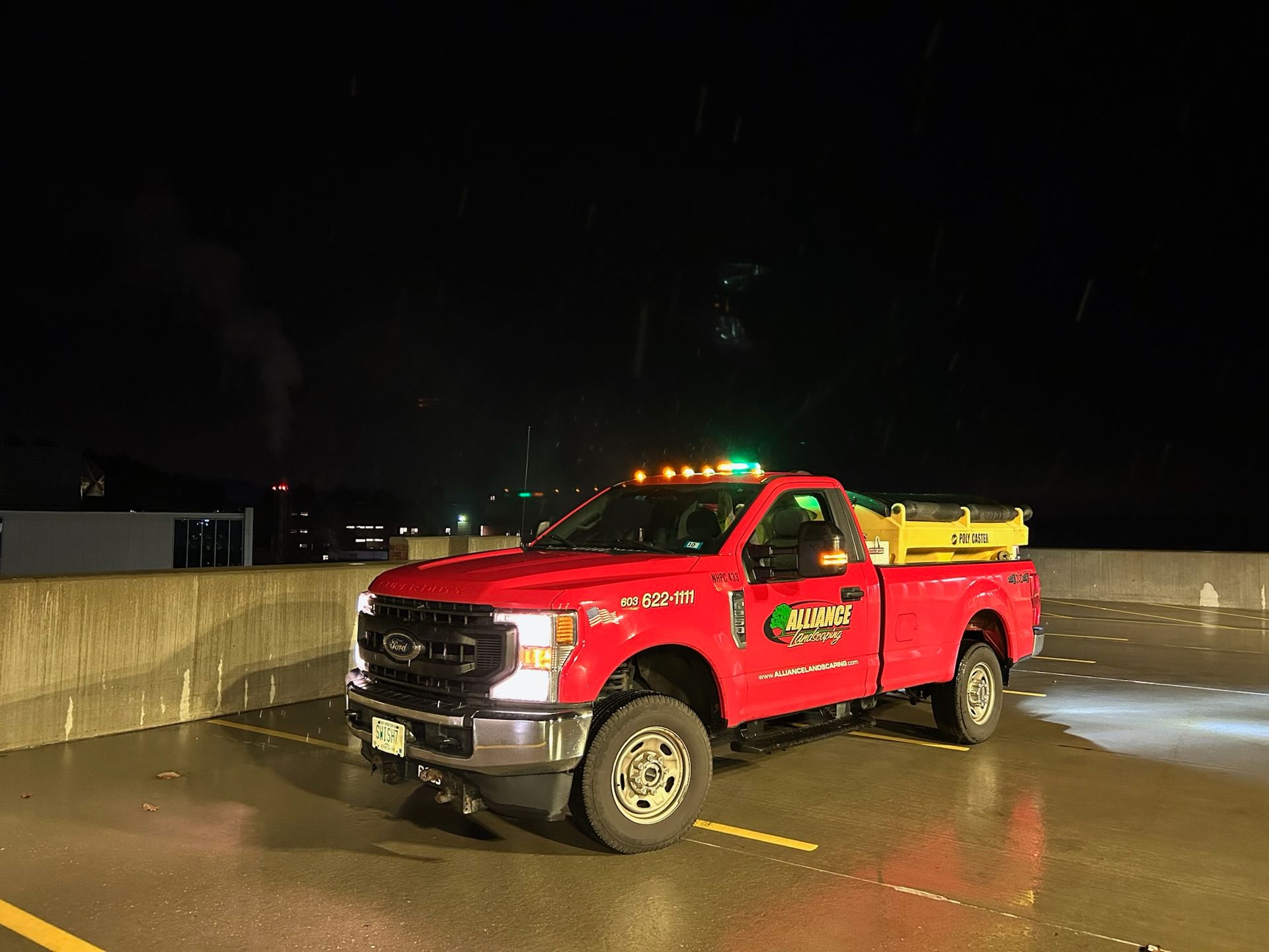 Red truck with yellow equipment on the back parked on a wet rooftop at night; green flashing lights.
