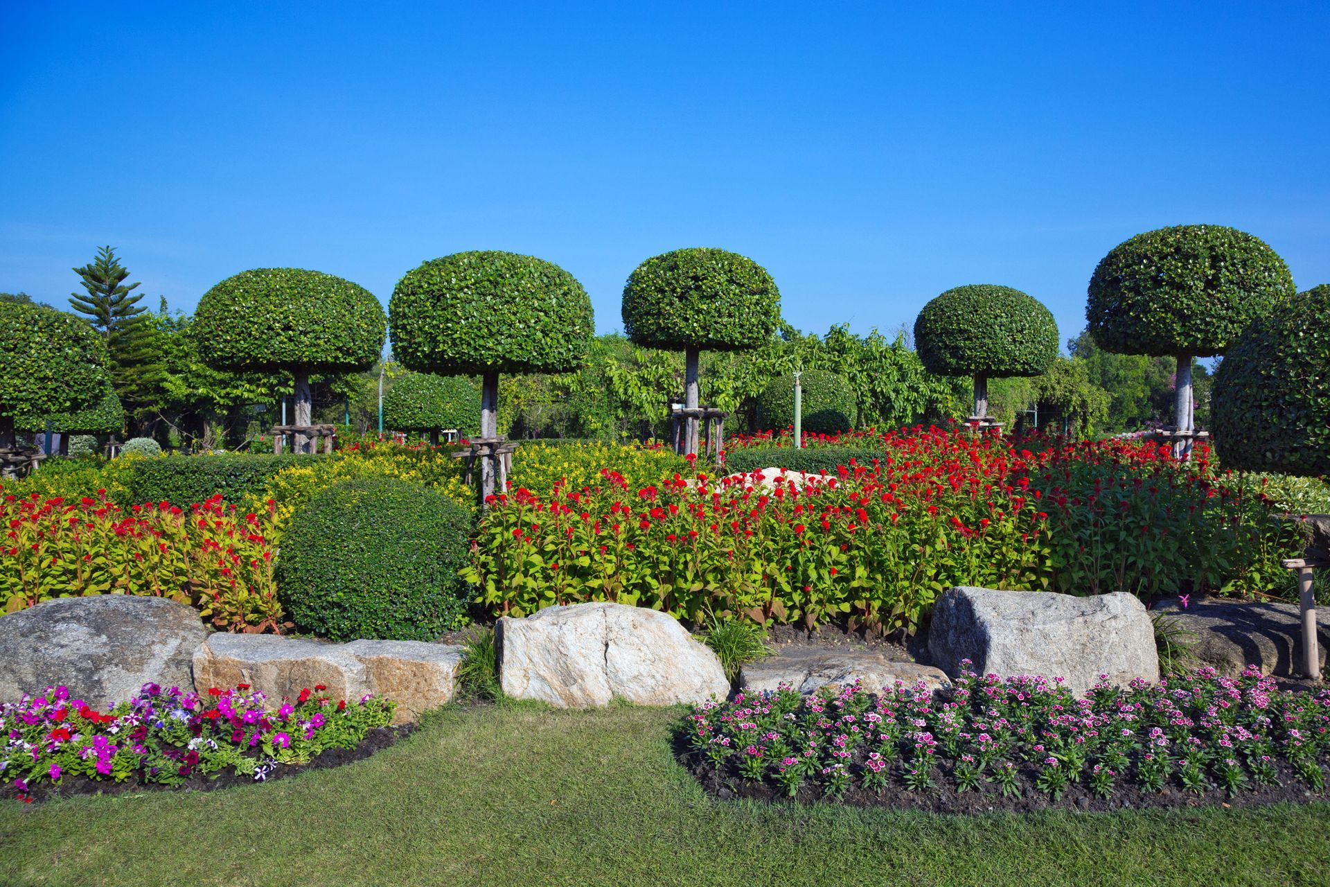 Colorful manicured garden with flower beds, trimmed round trees, rocks, and a bright blue sky