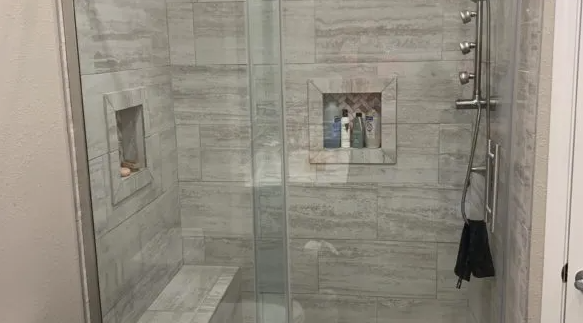 Shower stall with glass doors, tiled walls, built-in bench, and shower head with a black washcloth hanging.