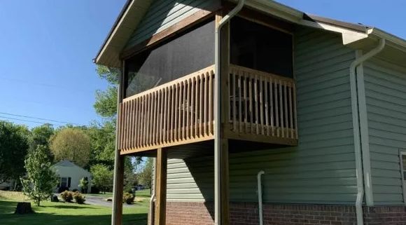 Wooden screened-in porch with railing, attached to a light green house with brick base, sunny day.