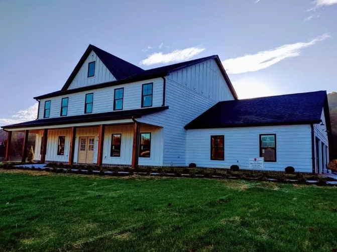 White farmhouse with dark roof and trim, wooden porch, set on a grassy hill.