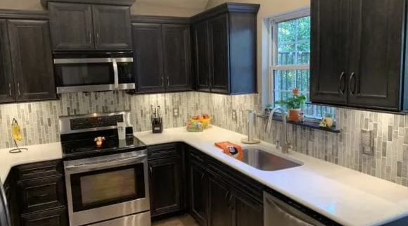 Dark kitchen with stainless steel appliances, white countertops, and mosaic backsplash.
