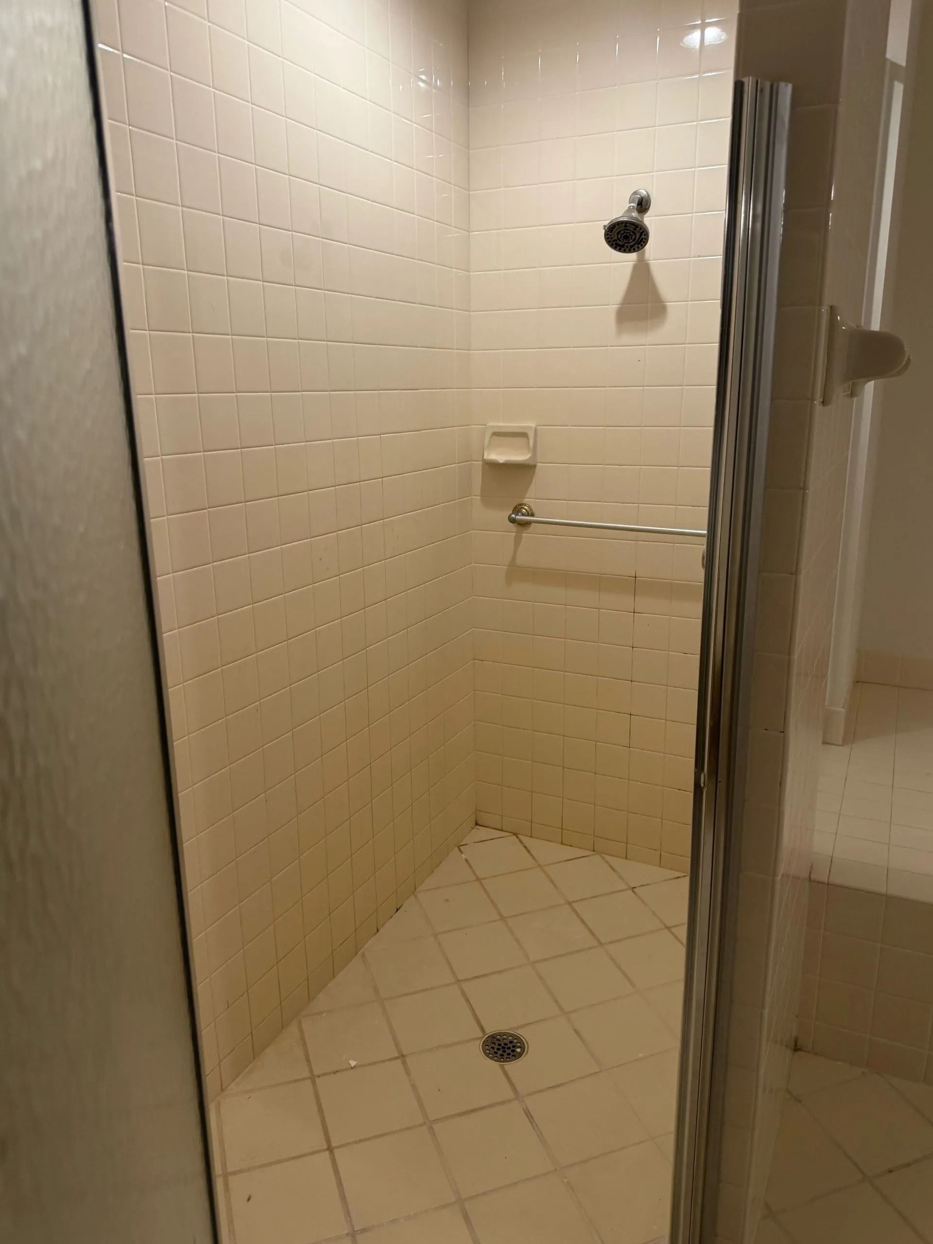 Shower stall with off-white tile walls and floor. A glass door is on the right. A showerhead and grab bar are visible.