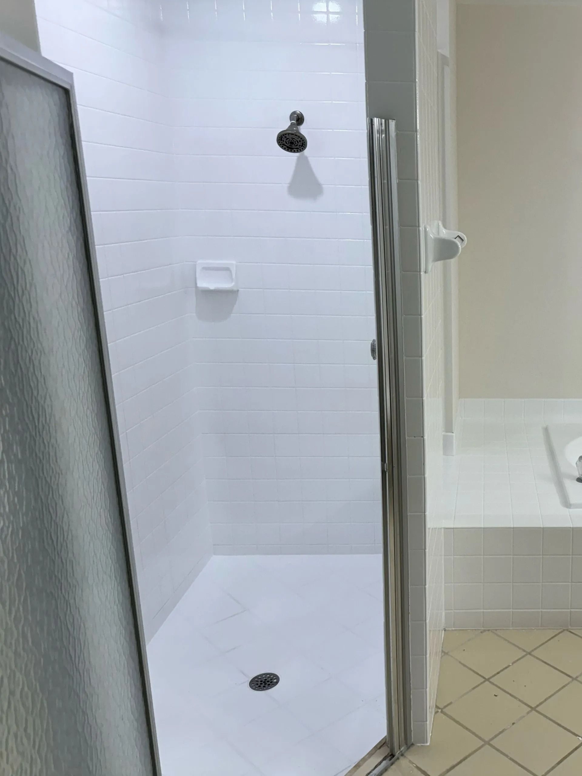 White tiled shower with glass door, showerhead, and built-in soap dish. Adjacent to a white tub.