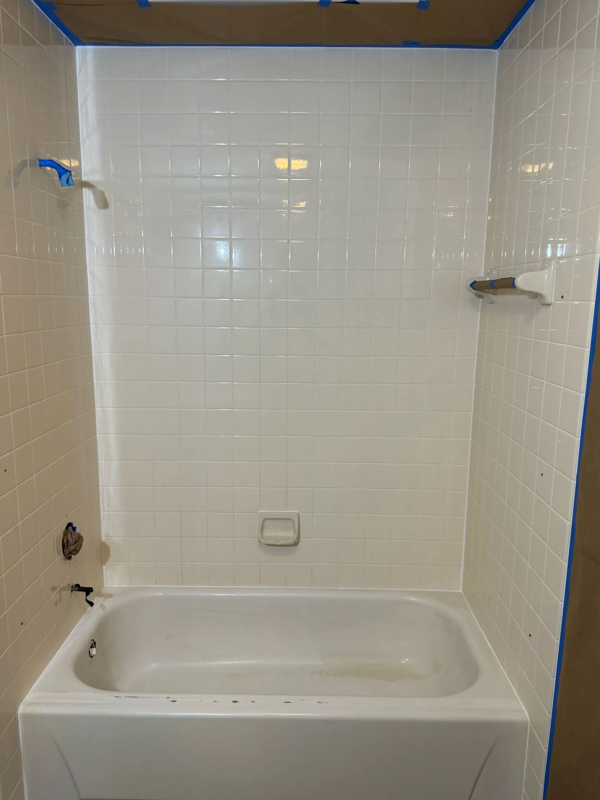 White-tiled shower enclosure with a bathtub, painted and taped. Faucet and soap dish visible.