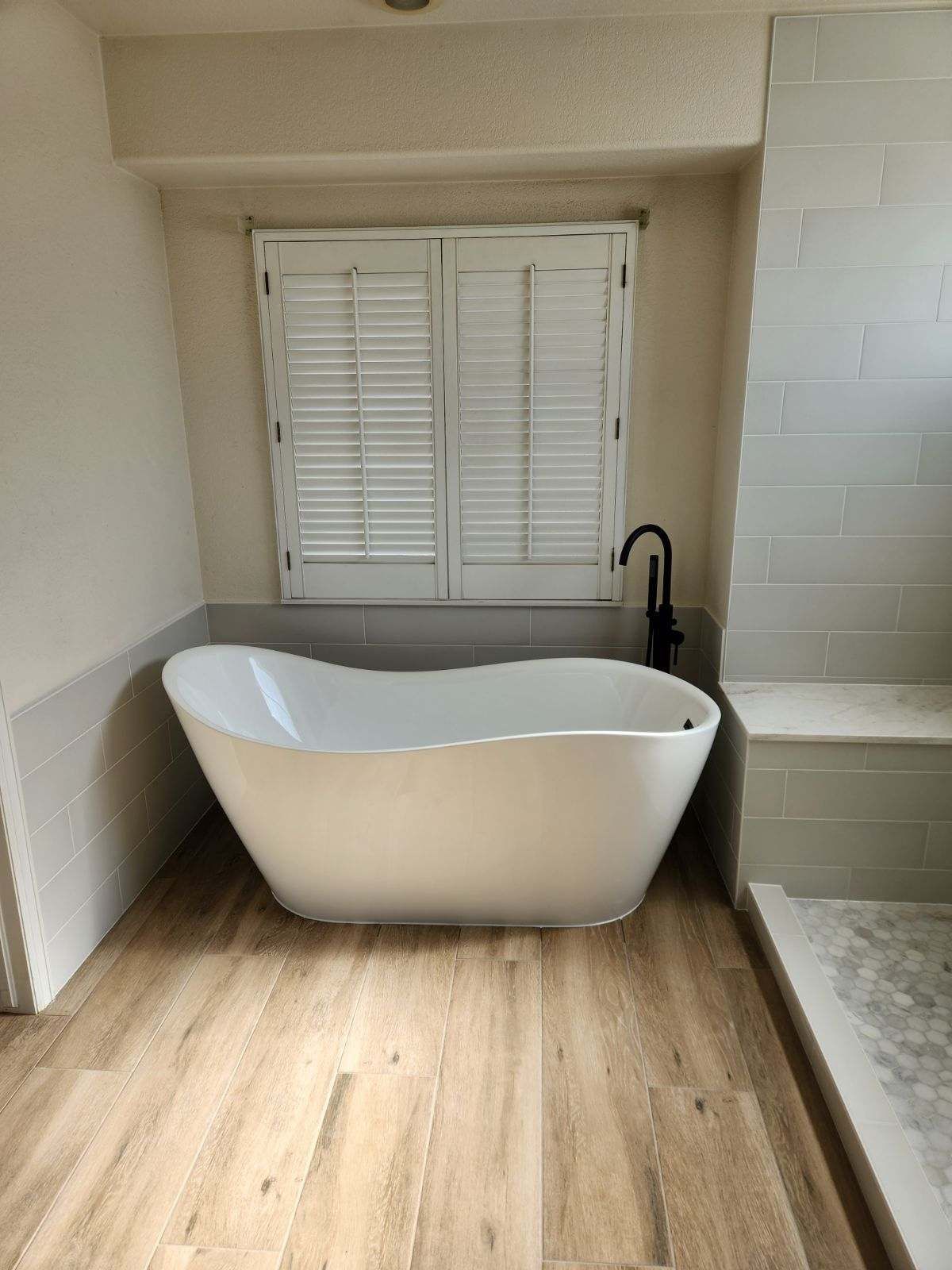 A white freestanding bathtub under a window with white shutters in a bathroom with wood-look tile flooring.